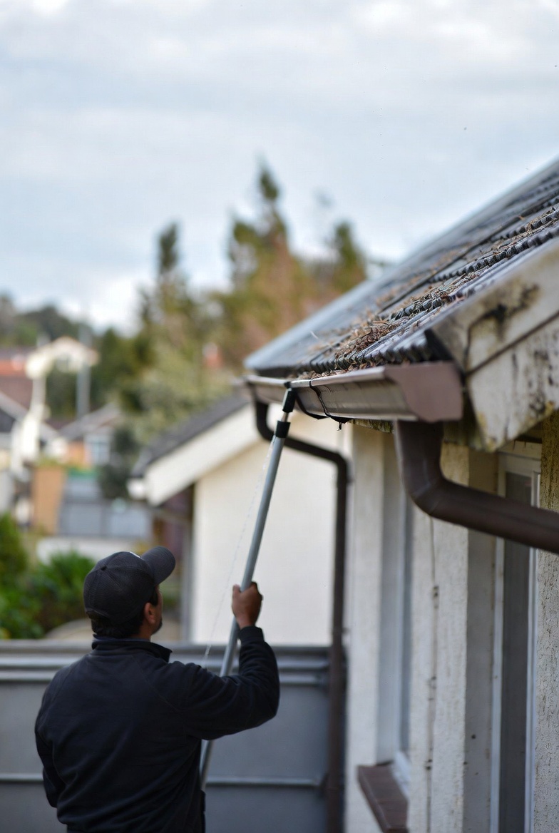 A gutter cleaning technician clearing leaves from a residential roofline while safety gear, ladder stabilizers, and cleanup tools sit below